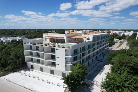 Aerial view of a modern multi-story residential building with white and grey facades under construction, surrounded by green trees and suburban landscape