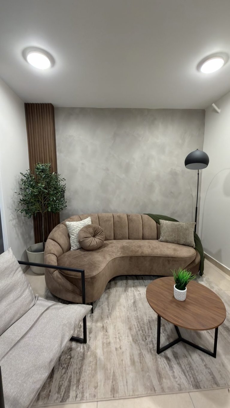Modern living room with curved beige sofa, wooden side table, potted plants, floor lamp, and recessed ceiling lights