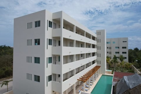 Modern white apartment complex with multiple balconies overlooking a turquoise pool and lounge area surrounded by tropical vegetation
