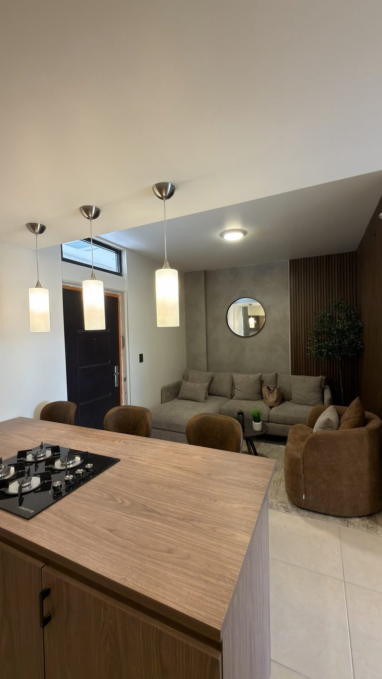 Modern open-plan kitchen and living room with wooden countertop, pendant lights, beige sofa, and dark entrance door