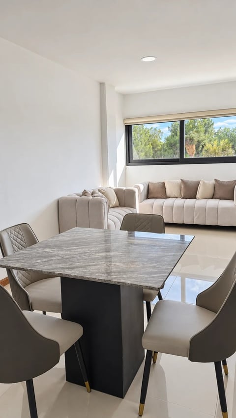 Sala comedor con mesa de comedor, sillas beige, sofá y ventanales con vista a los árboles