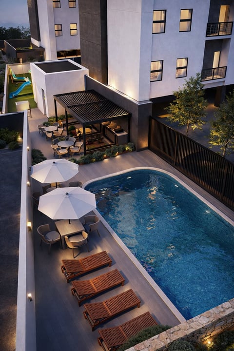 Patio residencial moderno con alberca rectangular, camastros de madera con sombrillas blancas, pérgola y edificio de departamentos al atardecer