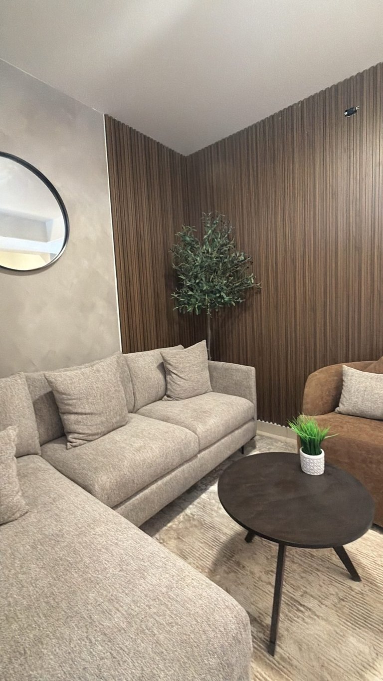 Modern living room with beige sectional sofa, black round table, wooden wall paneling, round mirror, and potted plant