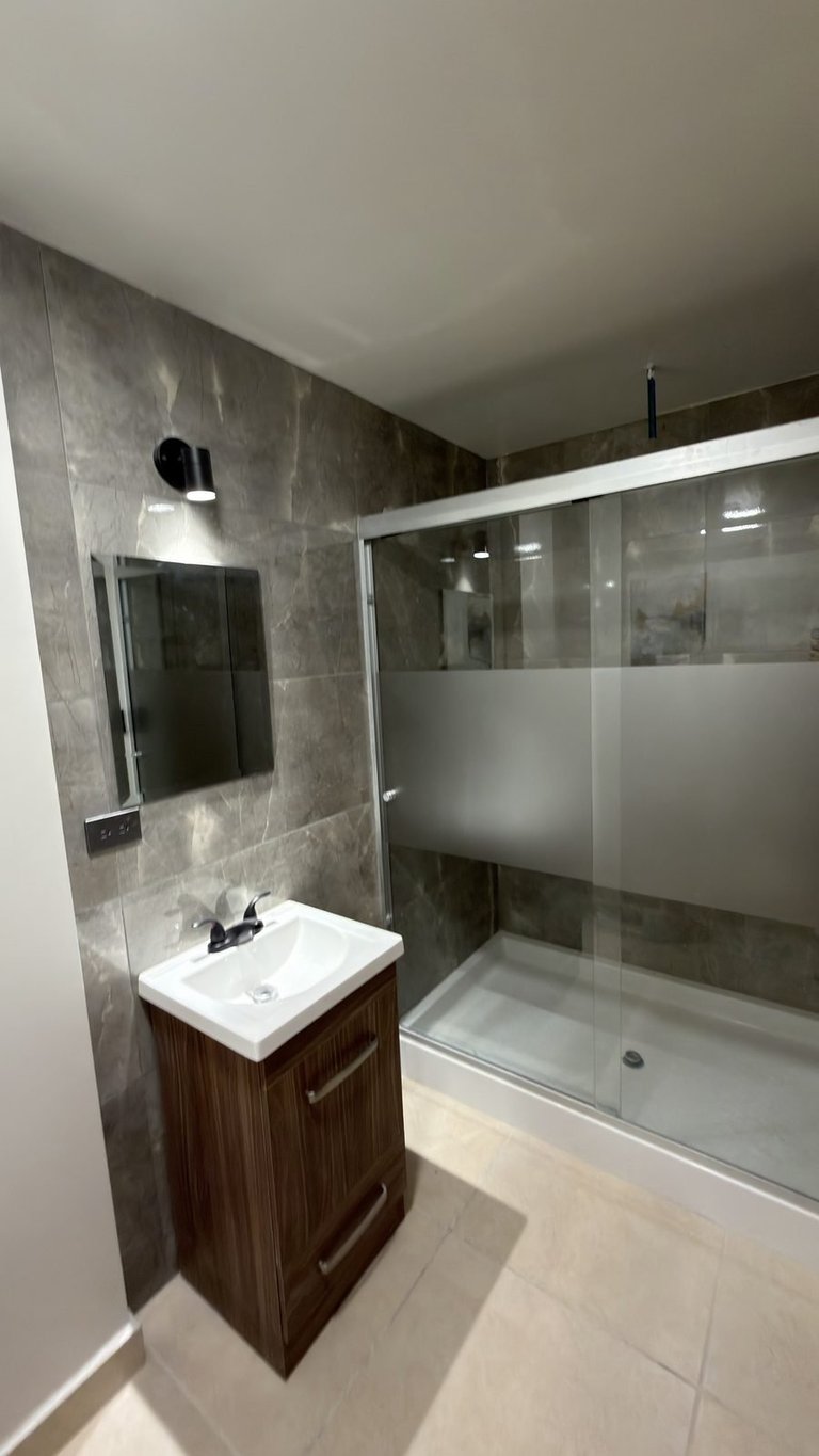 Modern bathroom with walk-in shower enclosure, wood vanity sink, gray tile walls, and frosted glass doors