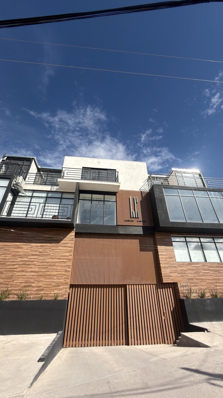 Fachada real de London Cantera: edificio residencial moderno de varios pisos con fachada de ladrillo y listones de madera bajo cielo azul con nubes blancas