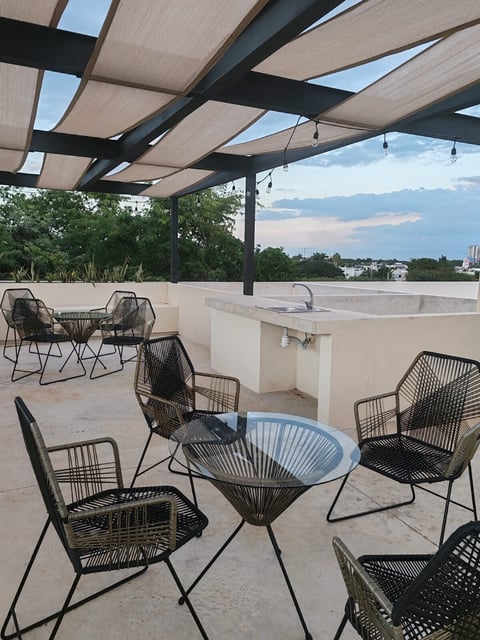 Modern rooftop terrace with black woven chairs, glass-top table, built-in hot tub, retractable shade panels, and city skyline view at dusk