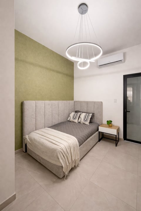 Recámara moderna con cama tapizada, pared de acento verde olivo, lámpara circular blanca y decoración minimalista