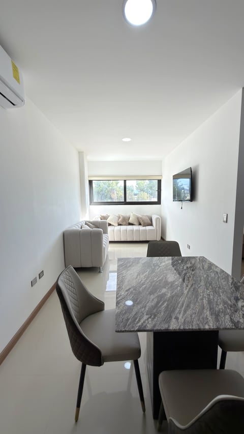 Estancia con mesa de comedor de mármol, sillas grises, sala blanca y televisión en espacio abierto