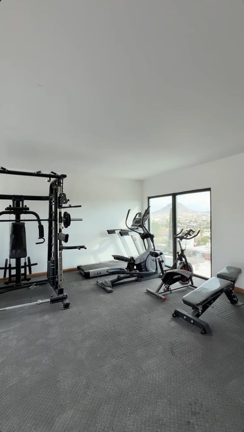Gimnasio equipado con bicicleta estática, caminadora y máquina de pesas, con ventanales hacia la ciudad