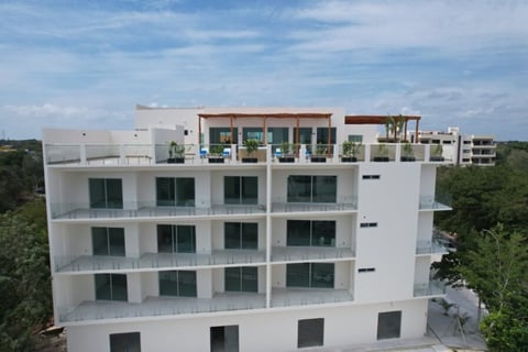 Modern white apartment building with four stories, large windows, and a rooftop terrace under blue sky surrounded by green trees