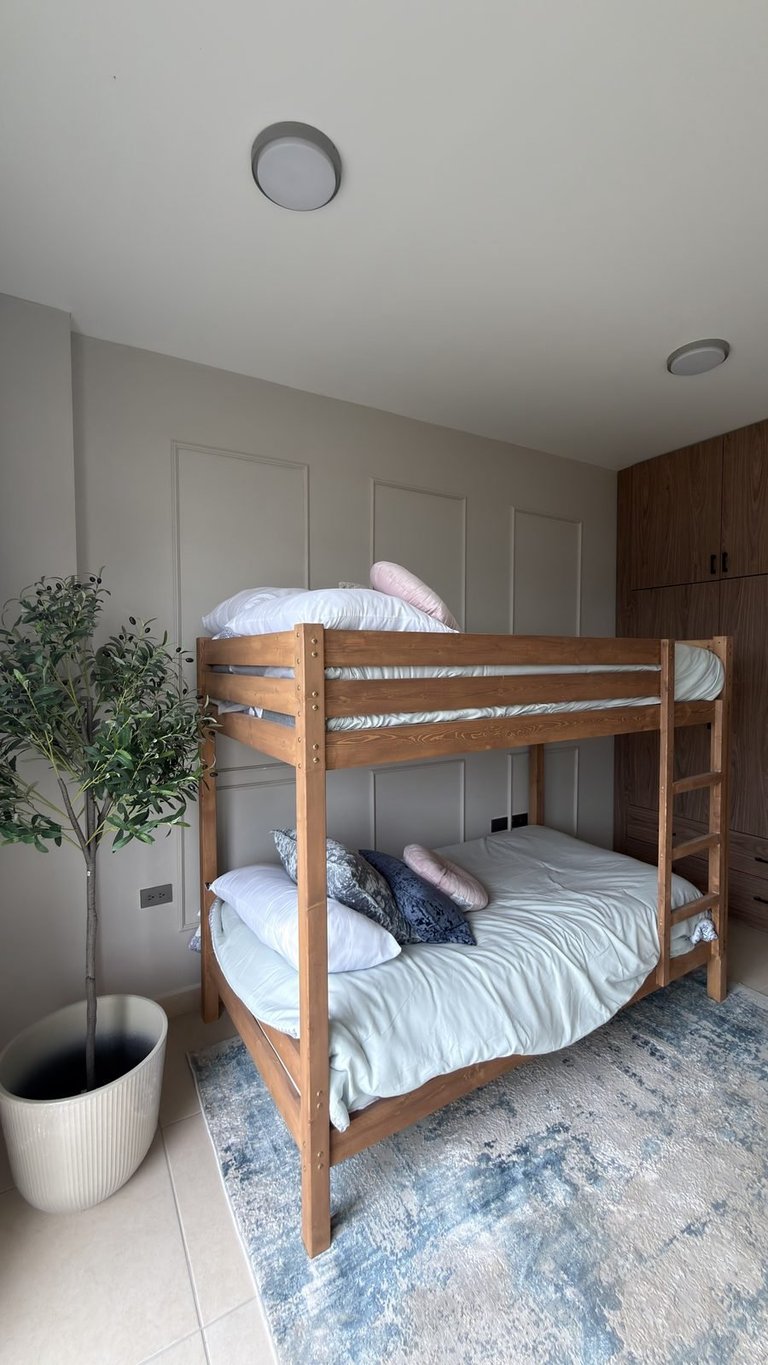 Wooden bunk bed with white bedding in a bedroom with gray walls, potted plant, and dark wood cabinetry